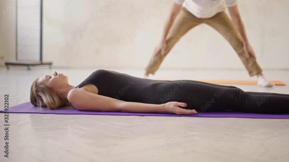 couple in white yoga studio. side view sporty relaxed woman practicing yoga in yoga class, lying in resting pose Savasana or Dead Body exercise, man stretches on background. Yoga Practice Exercise couple in white yoga studio. side view sporty relaxed woman practicing yoga in yoga class, lying in resting pose Savasana or Dead Body exercise, man stretches on background. Yoga Practice Exercise