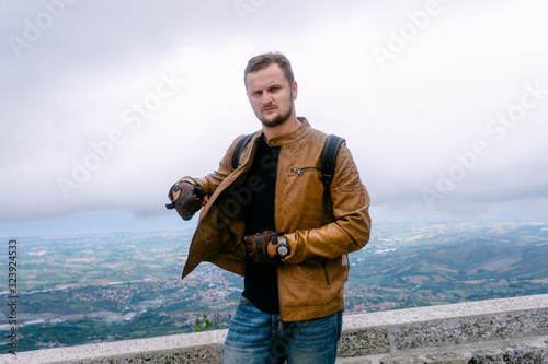 Portrait of a man dressed in a stylish brown leather jacket and jeans. backpack watches and biker gloves. posing on top of a mountain with a view of the city. San Marino, Italy