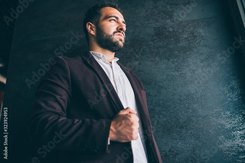 Serious caucasian prosperous male owner dressed in trendy expensive outfit looking away thoughtfully, handsome successful confident businessman posing near wall promotional background indoors.