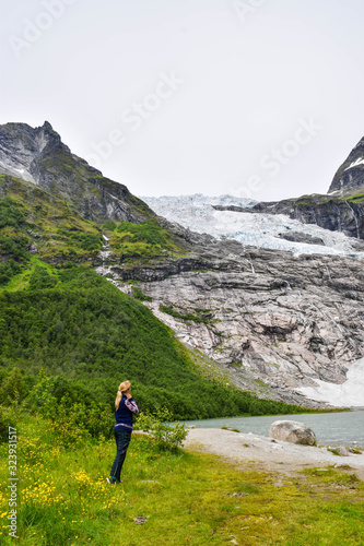 The young girl looks at the Boyabreen glacier, which is the sleeve of large Jostedalsbreen glacier and wild yellow flowers in the foreground. Melting glacier forms the lake with clear water. Norway. © jana_janina