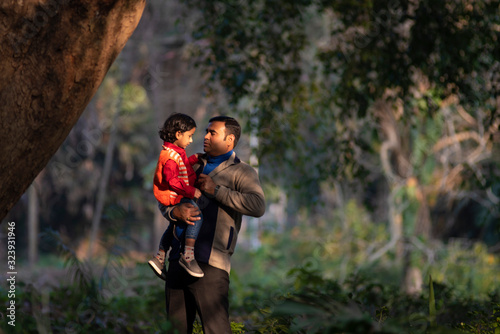 An Indian brunette father and his baby boy in winter garments enjoying themselves in winter afternoon in light and shadow in green forest background. Indian lifestyle and parenthood. © abir