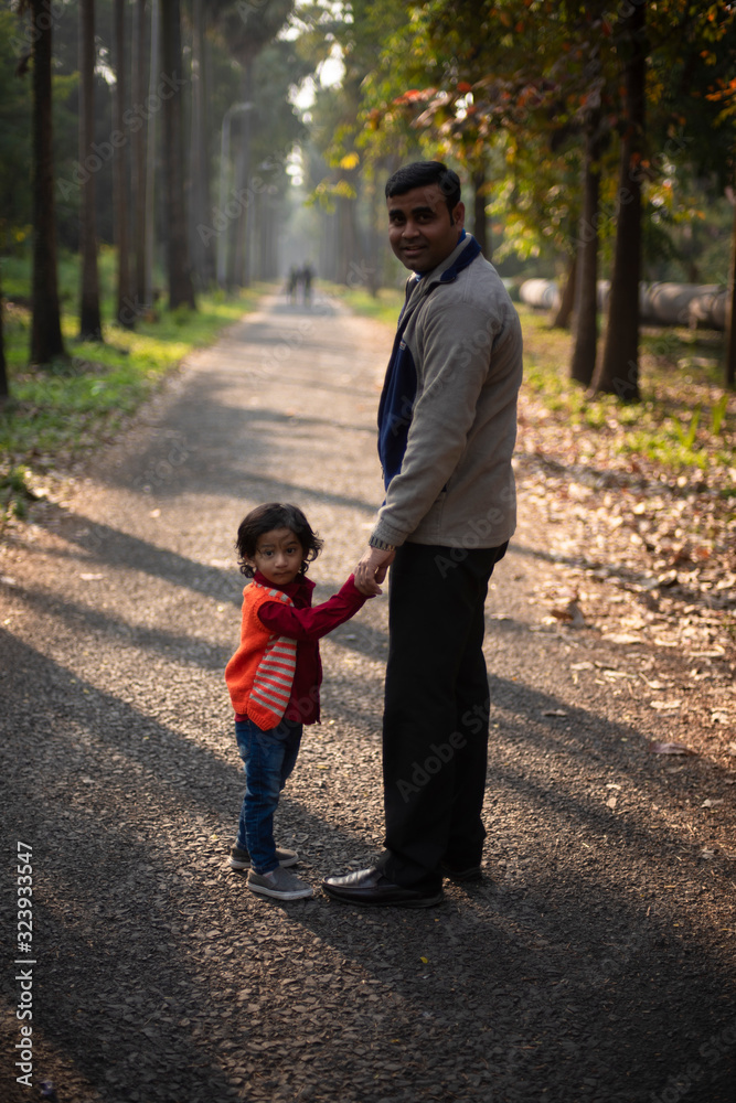 Fototapeta premium An Indian brunette father and his baby boy in winter garments standing on a forest path in winter afternoon in natural green background. Indian lifestyle and parenthood.