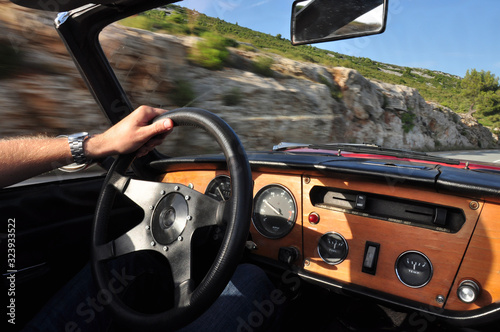 A man is driving an old red convertible classic car with one hand on the steering wheel. Dashboard shot from the passenger side with the feeling of speed visible thanks to the blur effect.  photo