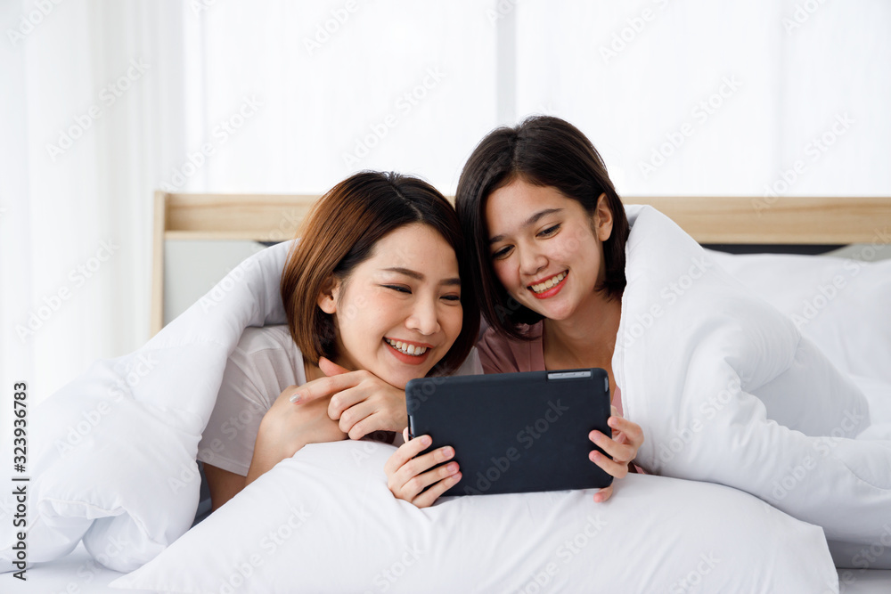 Two happy Asian women lie down on the bed and look at tablet. Concept for teenage or friends activity, lifestyle at home and romantic moments. Two happy Asian women lie down on the bed and look at tablet. Concept for teenage or friends activity, lifestyle at home and romantic moments.