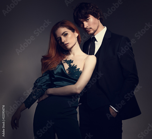 Sexy bright foxy hairstyle female woman in fashion green overalls dress posing with handsome man in black suit clothing on studio background. Closeup portrait