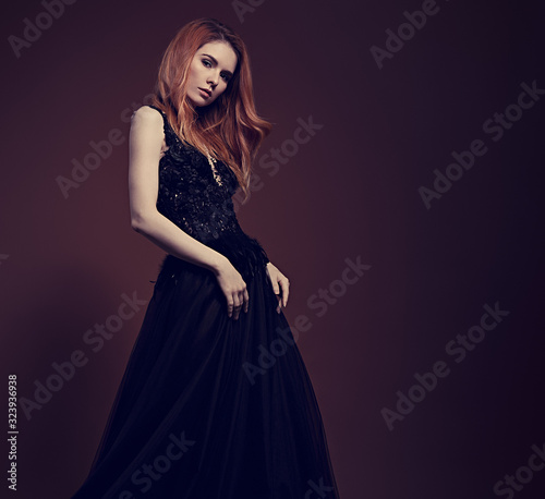 Beautiful redheaded young model with bright foxy hair posing in fashion chic black dress with long skirt on studio grey background. Mystical witch from fairy tale.