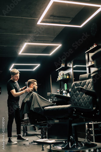 Salon for real men. Close-up photo of a young barber in a black t-shirt who is taking care of his customer’s hairstyle, using special tools.