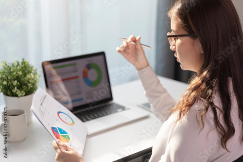 Young employee in eyeglasses working at office on laptop while sitting at table, Business woman analyze document in her hands, Graphs and diagram on notebook screen. © Suradech