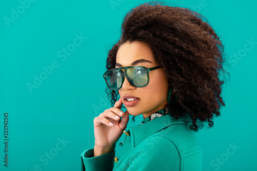 stylish african american woman in jacket looking at camera isolated on turquoise