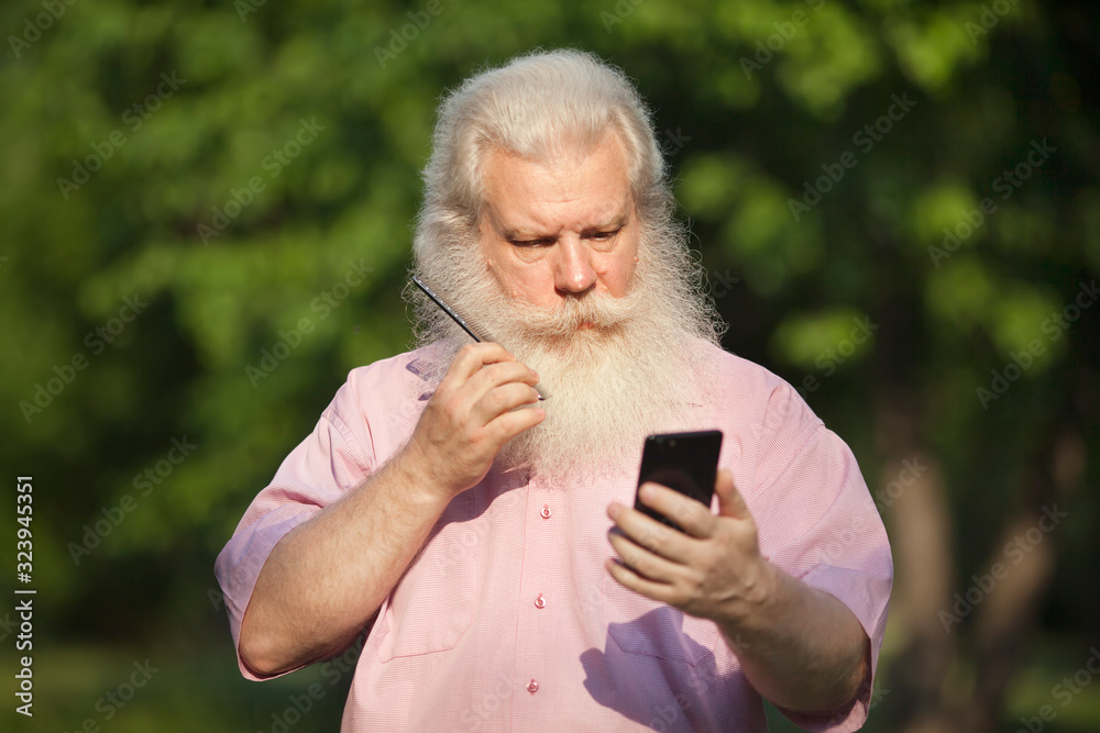 Serious stylish senior blond man in casual wear looking at smartphone as mirror and scratching his beard by comb. Closeup portrait of handsome person on sunlit green blur natural background. Serious stylish senior blond man in casual wear looking at smartphone as mirror and scratching his beard by comb. Closeup portrait of handsome person on sunlit green blur natural background.