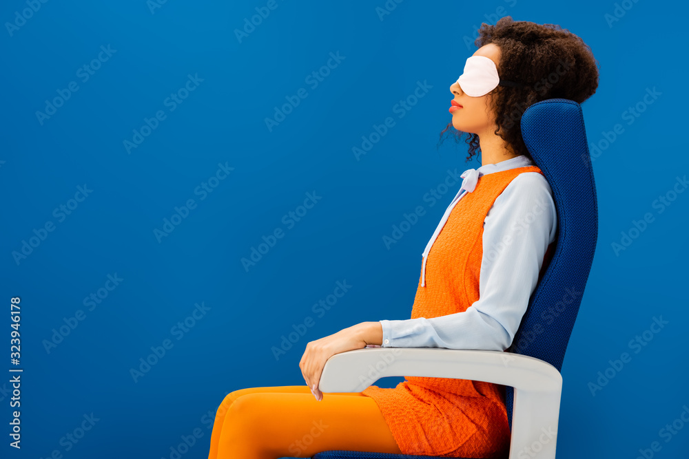 Fototapeta premium side view of african american with sleeping mask sitting on seat and sleeping isolated on blue