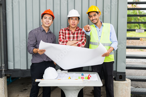 engineer and construction team holding blueprint on hand and pointing finger to building. they are checking construction process in construction site area.