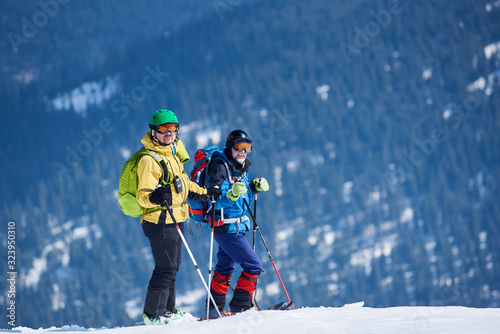 Two male skiers hikers in skiing equipment with backpacks on skis on background of mountain snowy slope covered with green spruce trees on frosty day. Winter tourism, ecological holidays concept.