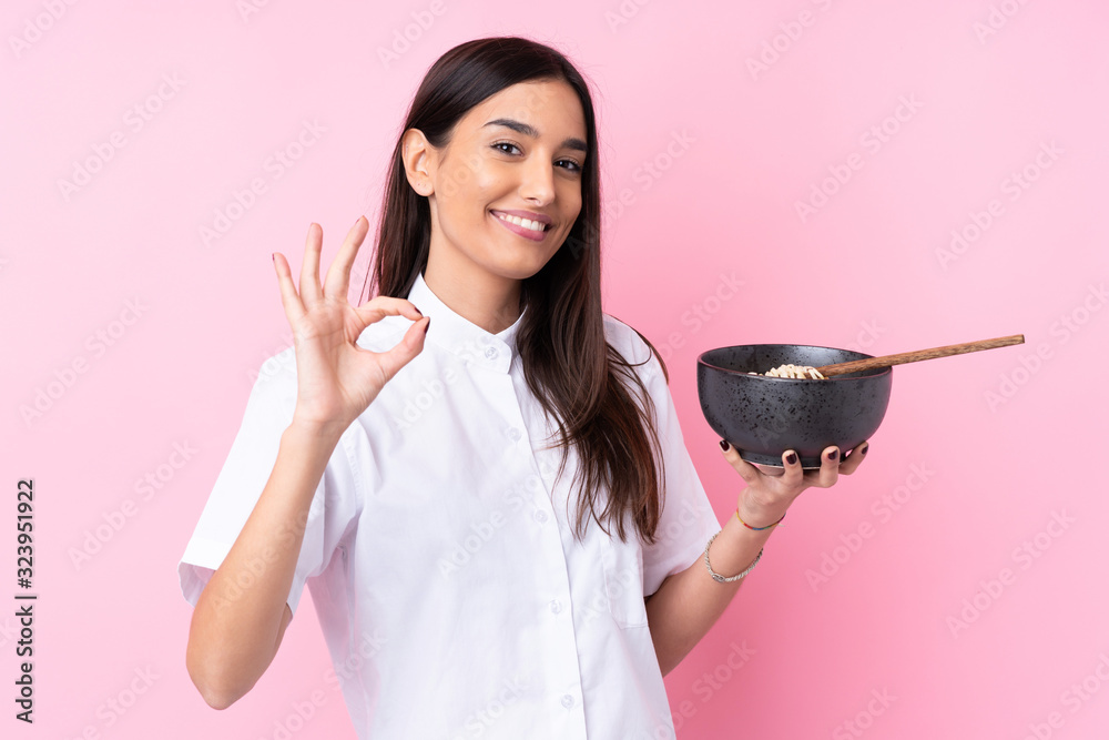 Young brunette woman over isolated pink background showing ok sign with fingers while holding a bowl of noodles with chopsticks Young brunette woman over isolated pink background showing ok sign with fingers while holding a bowl of noodles with chopsticks
