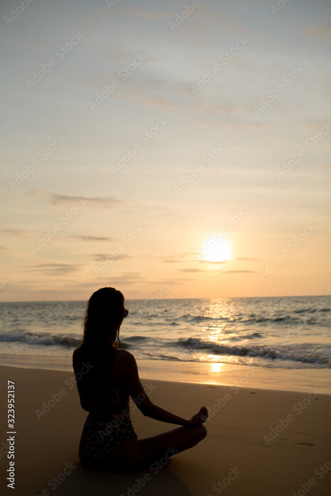a girl with glasses sits on the sand at sunset in the evening and meditates on the beach in Thailand and watches the sun set in a yoga pose