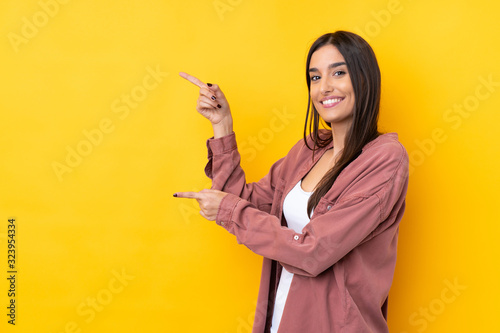 Young brunette woman over isolated yellow background pointing finger to the side and presenting a product photo