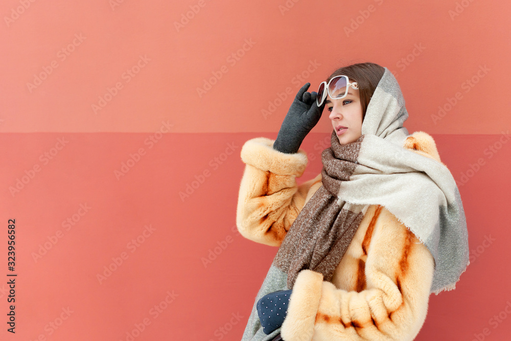 Fototapeta premium Fashionable portrait of a pretty young girl in a scarf in a fur coat, sunglasses and colorful clothes on a red background