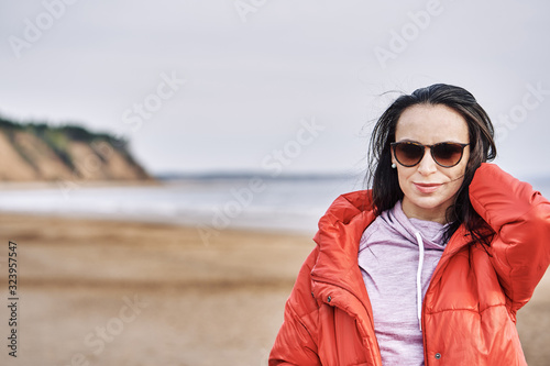 Portrait of a young brunette woman in sunglasses and a red jacket. Woman on a walk along the sandy shore of a large river on a spring day. Close-up. © afefelov68