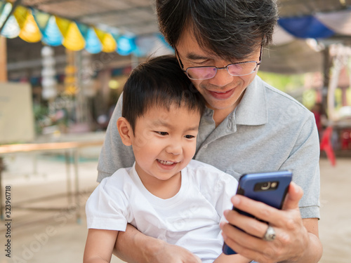 Asian little child boy watching smart phone with dad with happy smiling face. Kid and father looking to mobile phone together outdoor.