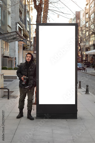 Young photographer stands in front of an empty billboard while taking pictures on the street