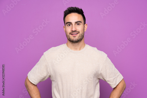 Young handsome man over isolated purple background posing with arms at hip and smiling