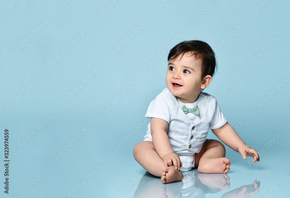 Baby boy in white bodysuit as a vest with bow-tie, barefoot. He smiling, sitting on the floor against blue background. Close up Baby boy in white bodysuit as a vest with bow-tie, barefoot. He smiling, sitting on the floor against blue background. Close up