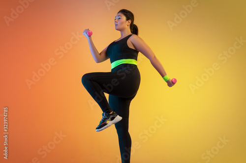 Young caucasian plus size female model's training on gradient orange background in neon light. Doing workout exercises with the weights. Concept of sport, healthy lifestyle, body positive, equality.