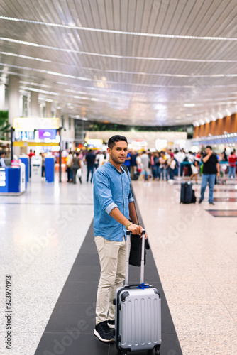 young latin traveller, with a big smile, ready to leave for Europe from Guayaquil, Ecuador, with grey suitcase and executive bag on his shoulders, in the terminal, people in the background