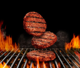Falling down beef cutlets for burgers. Meat roasted on metal barbecue BBQ grill with flaming fire and ember charcoal on black background. Close up