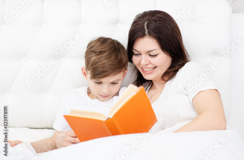 Mom and son are lying under the covers at home in the bedroom. Mom reads a book to her son