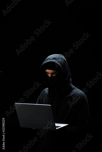 young hacker in hood using laptop isolated on black