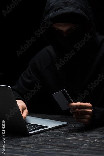 hacker in hood and mask using laptop while holding credit card isolated on black