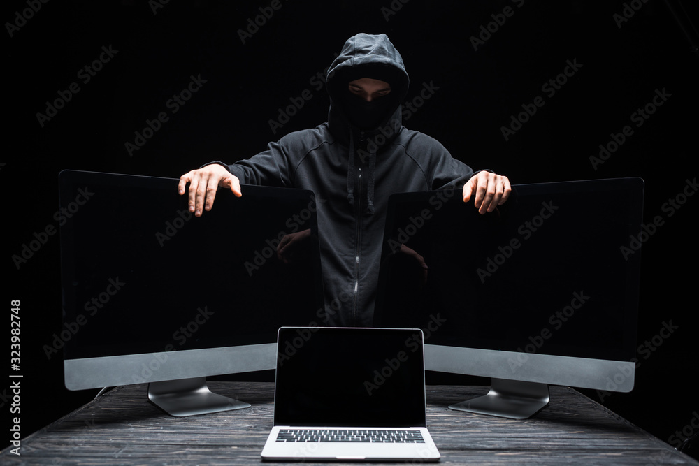 hacker in mask touching computer monitors near laptop with blank screen isolated on black hacker in mask touching computer monitors near laptop with blank screen isolated on black