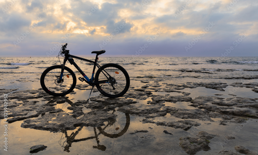 A biker has parked his mountain bike on the rocky coast near the Spanish port city of Torrevieja. It's morning after sunrise. A beautiful cloudy sky in the background.