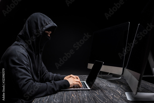 back view of hooded hacker with hands behind head near computer monitors with blank screen isolated on black