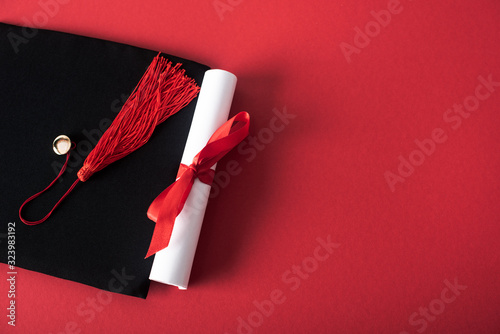 Top view of diploma with beautiful bow and graduation cap with tassel on red background photo