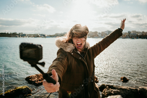 .Young tourist visiting a coastal town on the island of Malta. Taking pictures of herself with a small camera during a windy sunset wearing a coat. Lifesytle photo