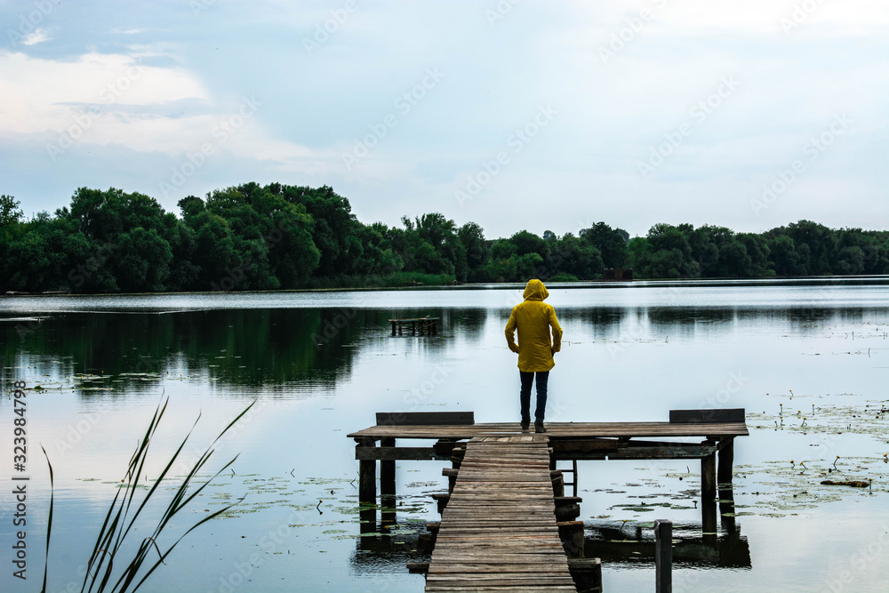 Stylish young man in yellow waterproof raincoat standing on the wooden pier by the lake or river