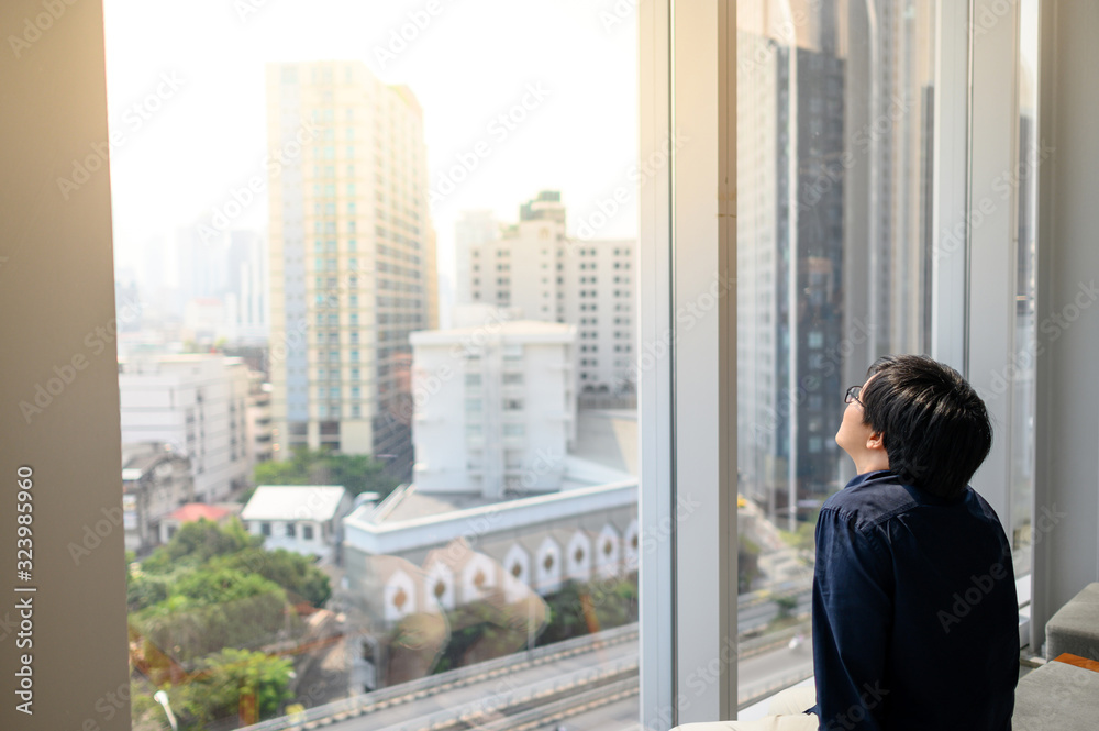 Young Asian business man sitting on bench in office building looking through the glass window with cityscape outside. Businessman relaxing during working in workplace. Work life balance concept Young Asian business man sitting on bench in office building looking through the glass window with cityscape outside. Businessman relaxing during working in workplace. Work life balance concept