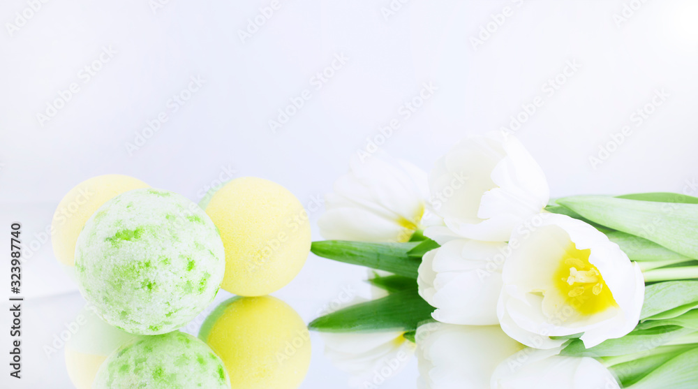 A green and yellow salt bomb on a white gray background is reflected, next to white tulips. Mineral makeup. Banner. Spa care. Banner. Copy space