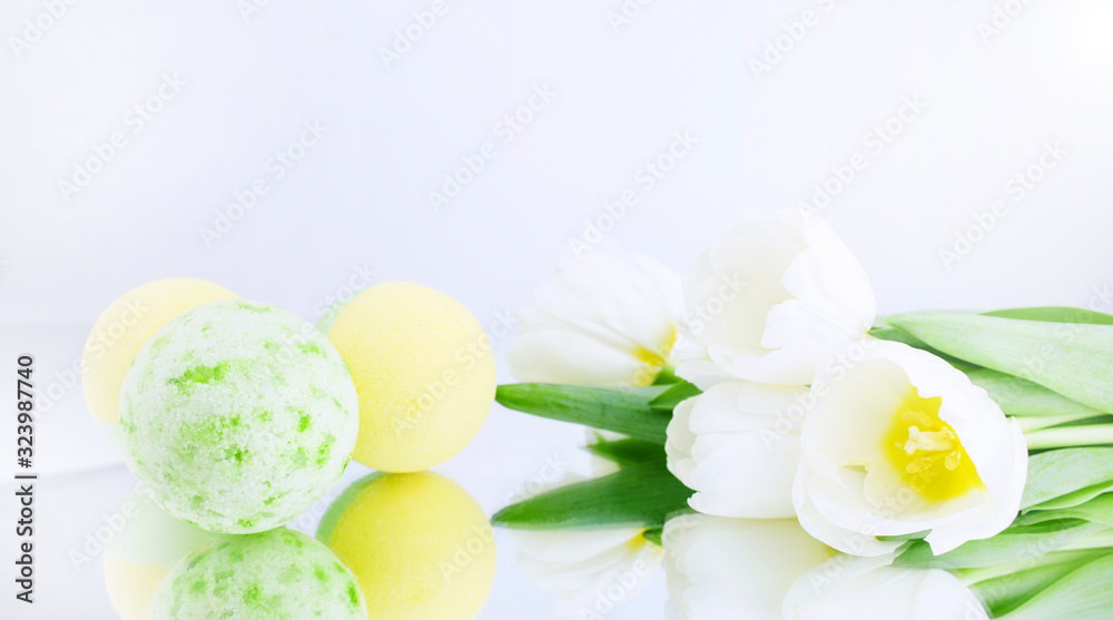 A green and yellow salt bomb on a white gray background is reflected, next to white tulips. Mineral makeup. Banner. Spa care. Banner. Copy space A green and yellow salt bomb on a white gray background is reflected, next to white tulips. Mineral makeup. Banner. Spa care. Banner. Copy space
