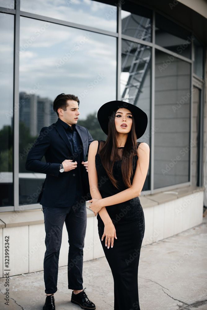 A beautiful, stylish pair of young people in black clothes and glasses stand against the background of an office building in the sunset. Fashion and style