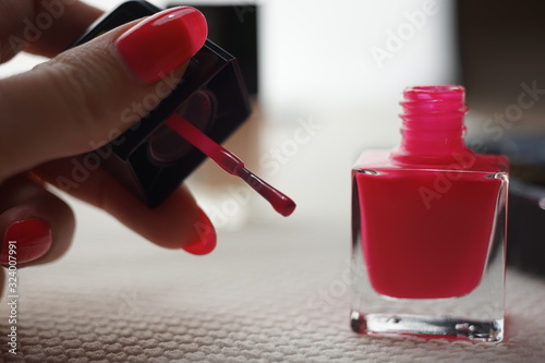 Open glass bottle with a brush and pink nail polish on a white table  female hand in blur