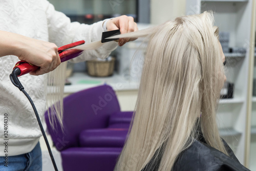 Straightening hair with flat iron. Blondie young woman in beauty salon. Hair procedures by professional hairdresser
