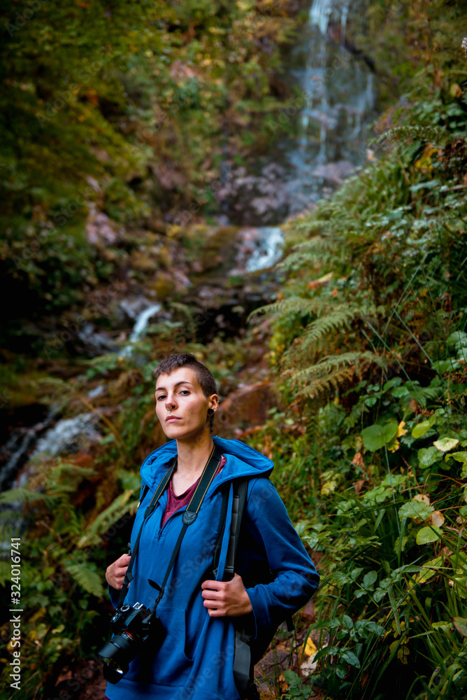 Focused young woman with professional camera and backpack looking at camera while standing at waterfall in forest in summer day Focused young woman with professional camera and backpack looking at camera while standing at waterfall in forest in summer day