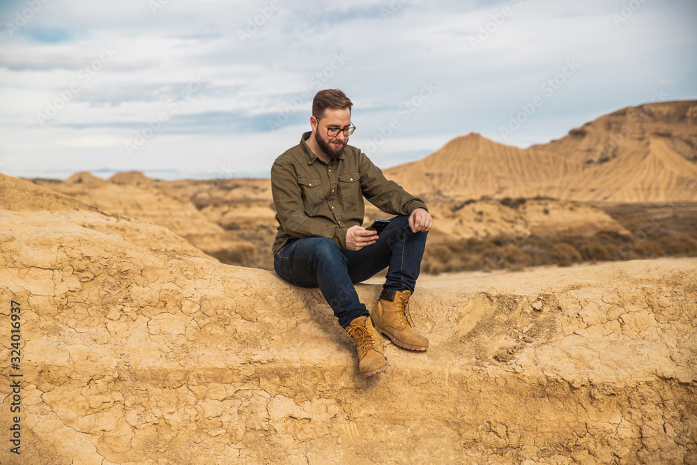 Content bearded male in casual wear and sunglasses using mobile phone while resting at foot of brown mountain with blue sky on background at Bardenas Reales Content bearded male in casual wear and sunglasses using mobile phone while resting at foot of brown mountain with blue sky on background at Bardenas Reales