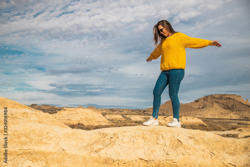 Side view of young female tourist in yellow sweater blue jeans and white sneakers spreading arms for balance while walking along peak of brown hill in Bardenas Reales, Navarre, Spain Side view of young female tourist in yellow sweater blue jeans and white sneakers spreading arms for balance while walking along peak of brown hill in Bardenas Reales, Navarre, Spain