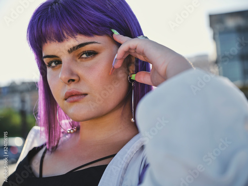 Fashion stylish woman with purple hairstyle touching face and leaning on metal fence in city center and confidently looking at camera in bright day photo