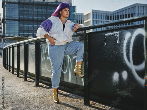 Fashion stylish woman with purple hairstyle leaning on metal fence in city center and confidently looking away in bright day photo