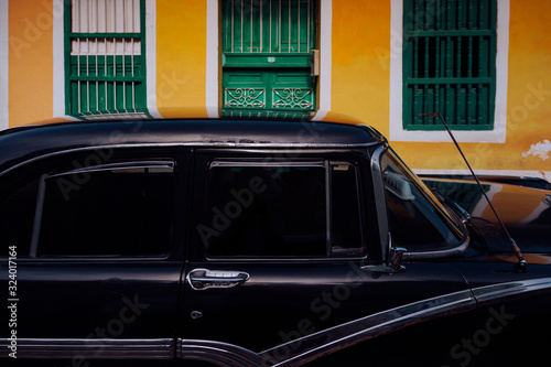 Small street with black vintage car in roadside between historical colorful buildings with bars on windows in Cuba